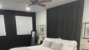 Ein modernes Schlafzimmer mit einem Bett, Nachttischen, einem Deckenventilator und einem großen Fenster mit Jalousien.