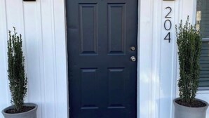 Eine schwarze Haustür mit Fußmatte, auf der steht: "Seien Sie unser Gast." Auf jeder Seite der Tür befinden sich je ein Blumentopf, und die Hausnummer 204 ist rechts an der Tür erkennbar.
