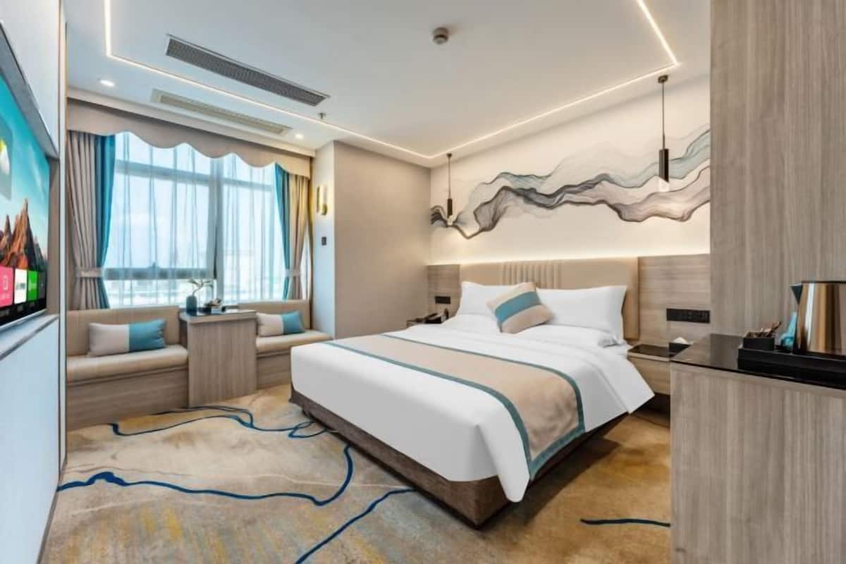A modern hotel room with a large bed, a seating area with a sofa and armchairs, a desk, and a view of the city.