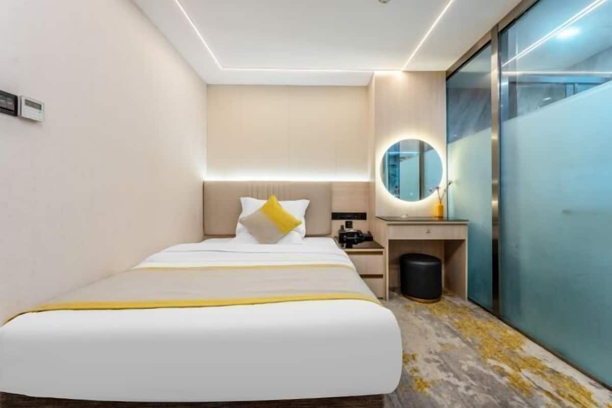 A hotel room with a bed, bedside table, mirror, and a trash bin.