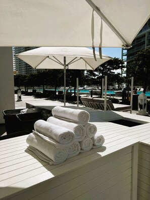 A poolside area with a stack of white towels, lounge chairs, and a large umbrella providing shade.