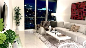A modern living room with a sofa, a coffee table, and a TV. There is a large window with a city view.