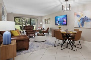 A modern living room with a brown leather sofa, a dining table with chairs, a coffee table, and a flat-screen TV mounted on the wall.