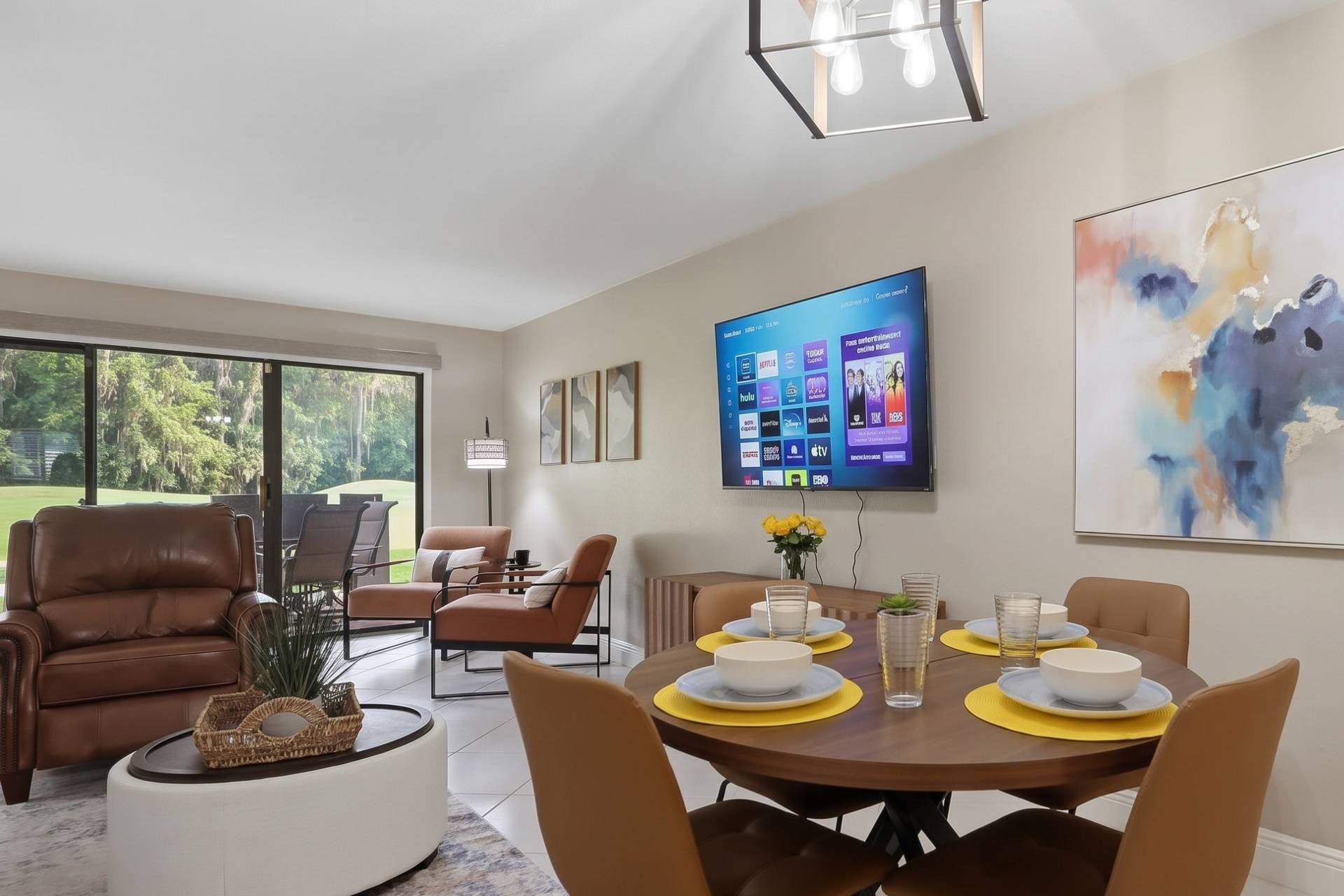A dining area with a round table set for four, surrounded by chairs, a television mounted on the wall, and a painting above the table.