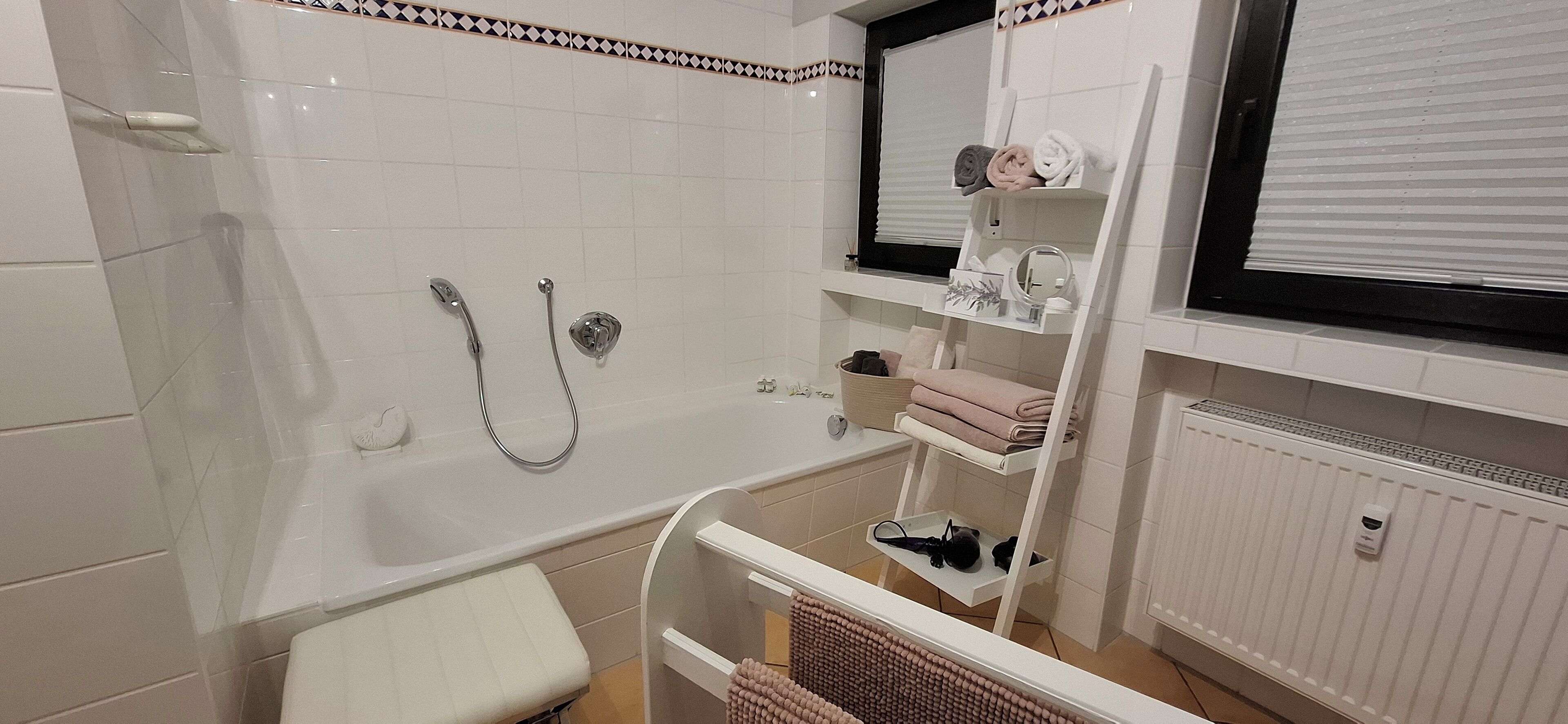 A bathroom with a bathtub, a shower, a toilet, and a sink. There are shelves with towels and toiletries.