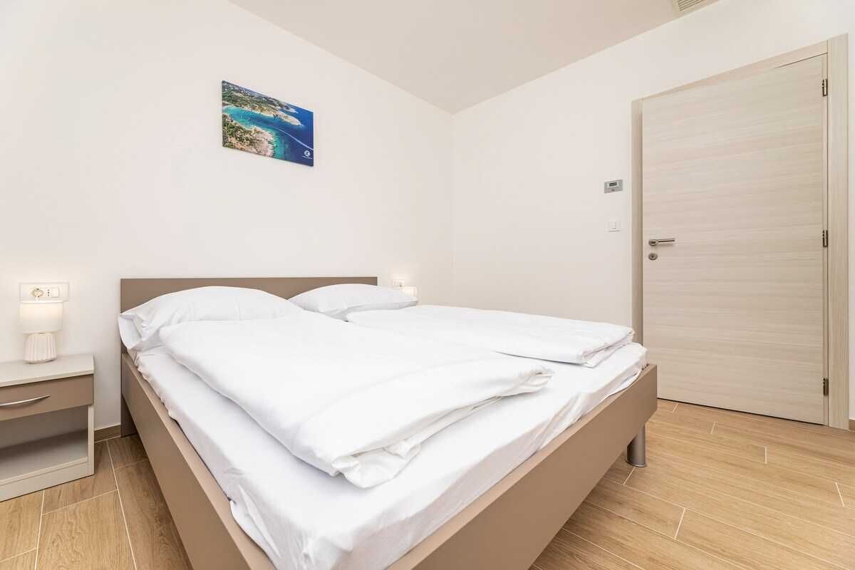 A neatly made bed with white linens in a room with a wooden floor, a nightstand with a lamp, and a door to the right.