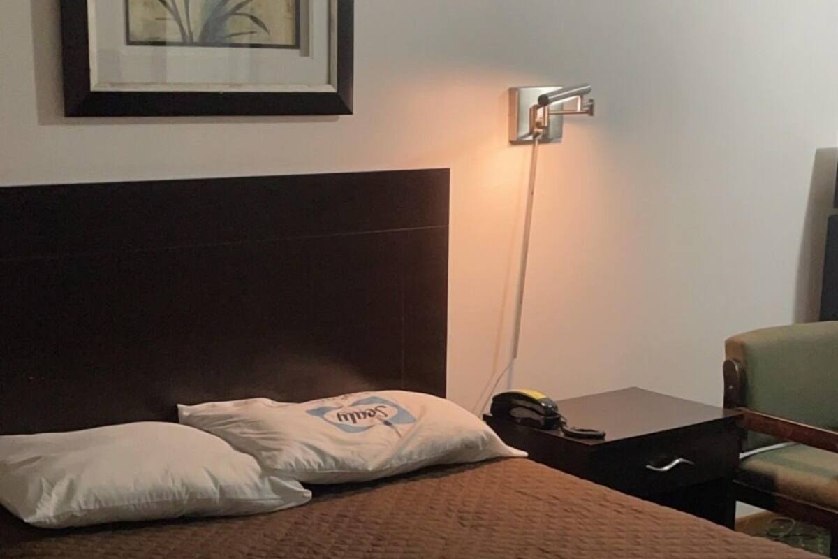 A hotel room with a bed, pillows, a nightstand, a lamp, a framed picture on the wall, and a chair.