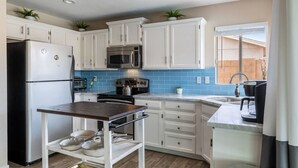 Una cocina moderna con armarios blancos, un refrigerador de acero inoxidable, un microondas y una estufa. Hay una pequeña mesa con vajilla y una tetera.