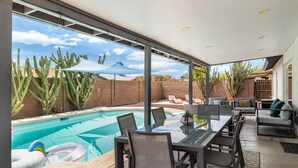Un área de piscina moderna al aire libre con una mesa y sillas de cristal, rodeada de cactus y un cielo despejado.