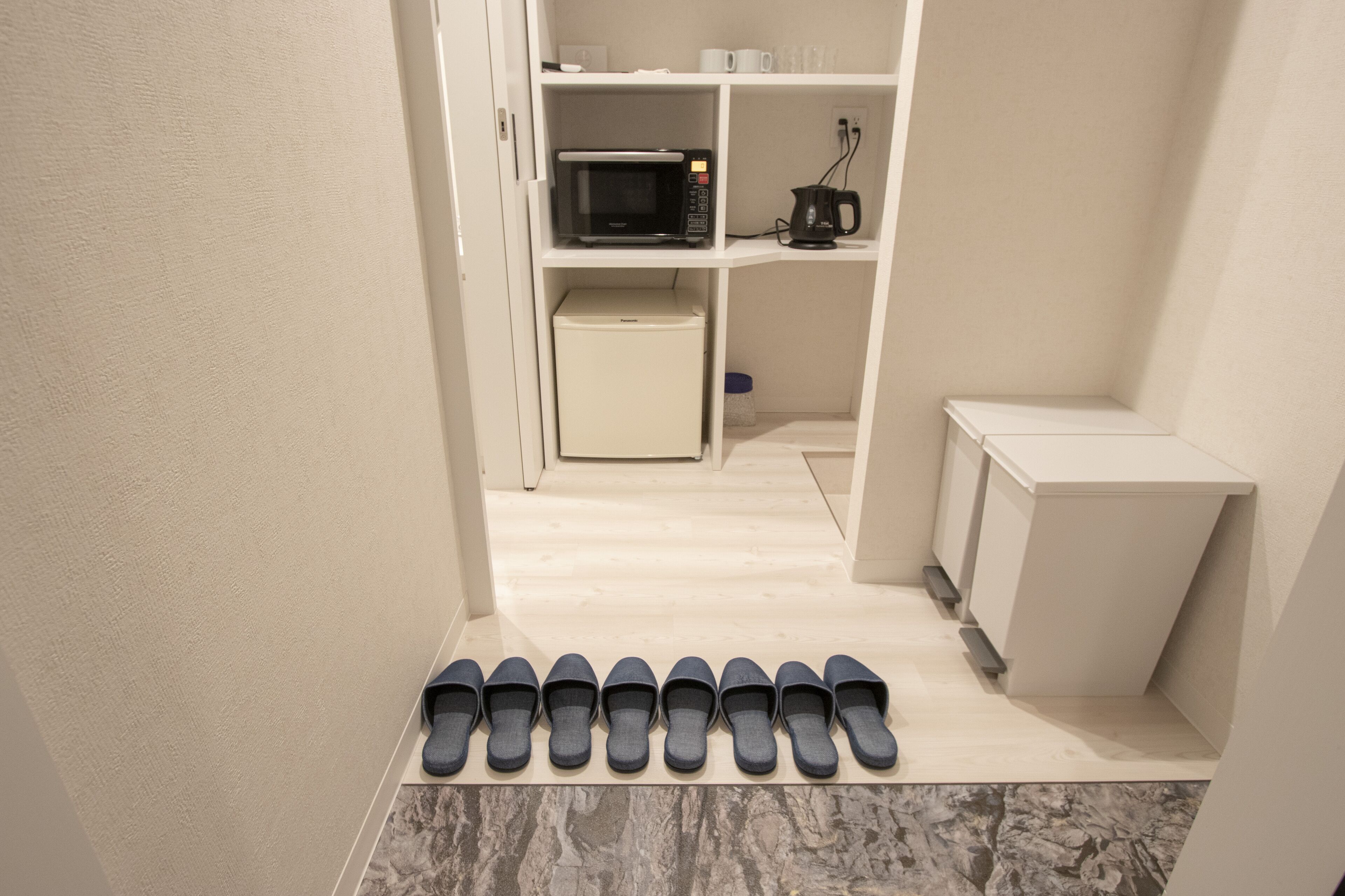 A hallway with a row of black slippers on a tiled floor, leading to a kitchen area with a microwave and a coffee maker.