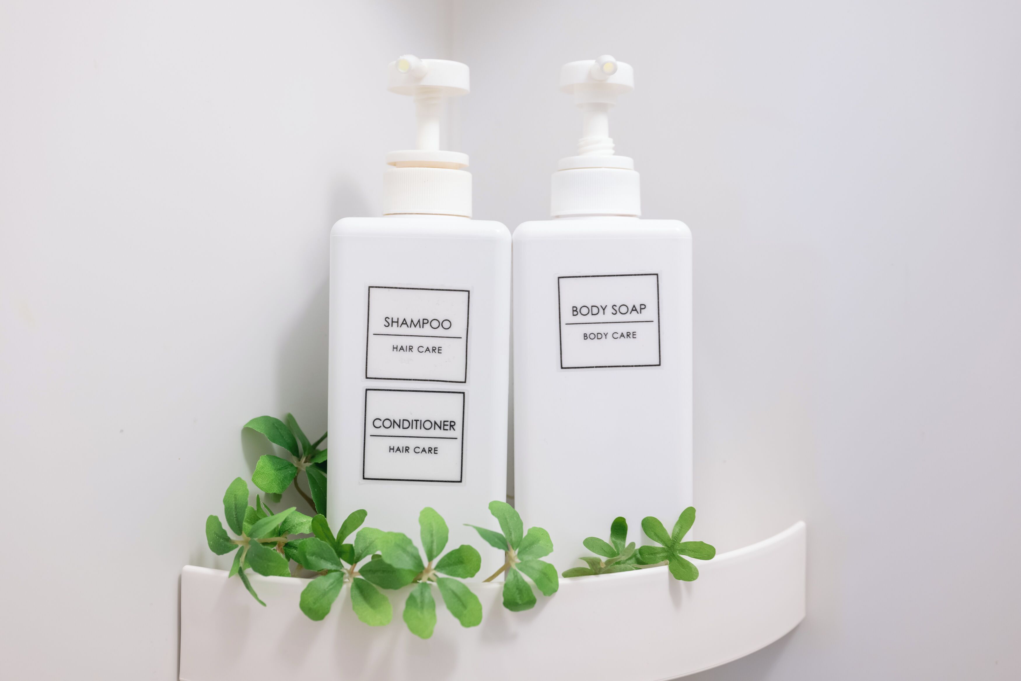 Dos botellas blancas de champú y gel de baño con dosificadores, etiquetadas para el cuidado del cabello y del cuerpo, sobre un estante blanco con hojas verdes.