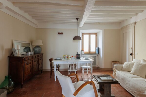 A dining area with a white table, chairs, and a window. There is a wooden cabinet with a mirror and a lamp on it. A white sofa is visible in the foreground. - City / Village Apartment in Cortona with 2 bedrooms sleeps 4 (Cortona)