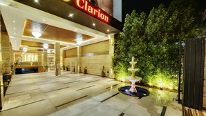 Entrada de hotel com balcão de recepção, uma fonte e área externa bem iluminada.