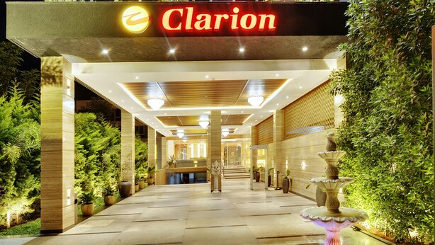 Entrada de hotel com uma fonte, plantas em vasos e uma placa com a inscrição 'Clarion'.