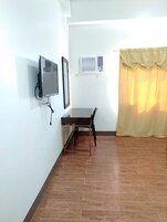 A room with a wooden floor, a table with chairs, a television, an air conditioning unit, and a curtain.