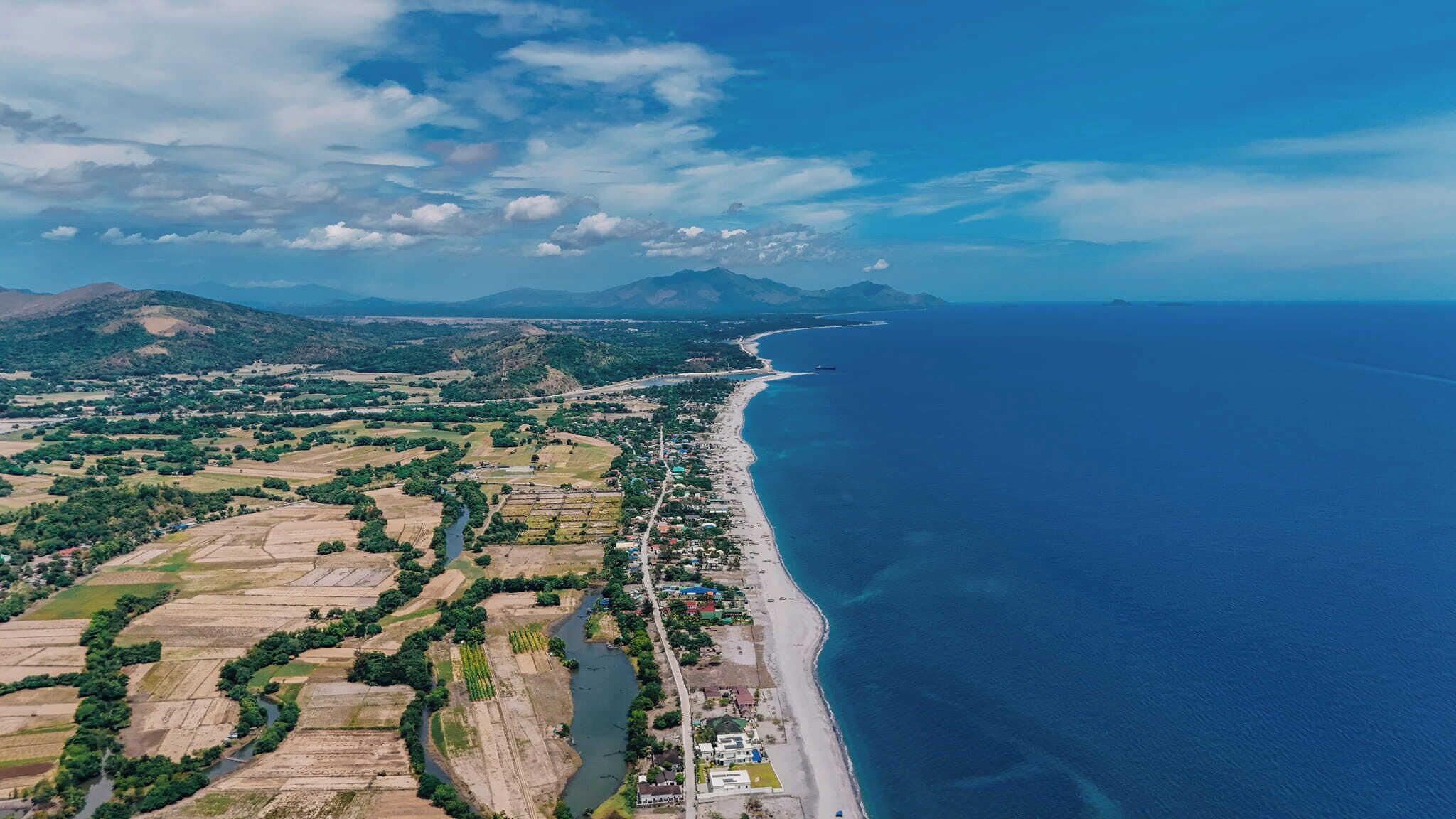 Vista aérea de uma área costeira com praia, campos agrícolas e uma estrada ao longo da costa.