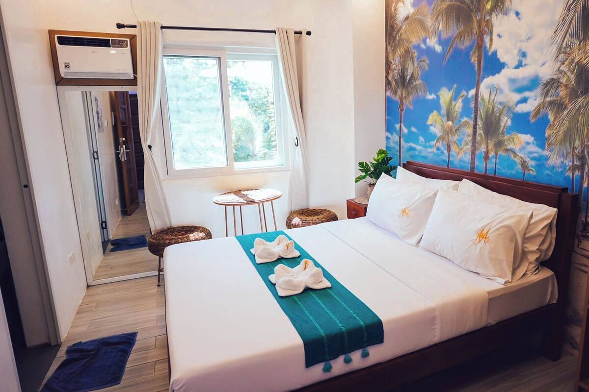 Habitación de hotel con cama, ventana con cortinas, aire acondicionado de pared y un mural tropical.