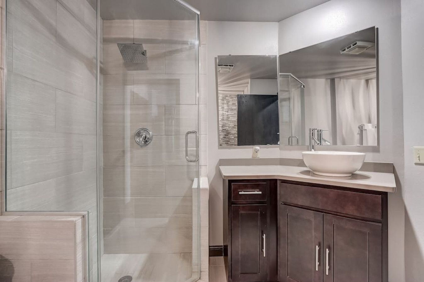 Un baño moderno con ducha de vidrio, un espejo grande y un tocador de madera oscura con lavamanos blanco.