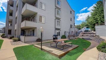 A multi-story apartment building with a well-maintained lawn, a picnic table, and a barbecue grill in the front yard.