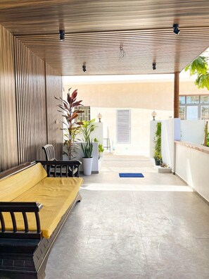 A covered outdoor seating area with a sofa and chairs, potted plants, and a view of the building in the background.