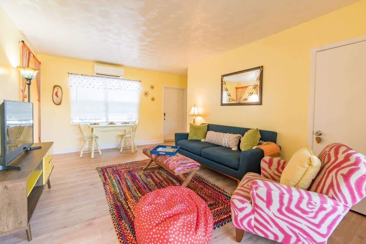Een woonkamer met een bank, fauteuil, hocker, een televisie, een bureau met stoelen en een raam met gordijnen.