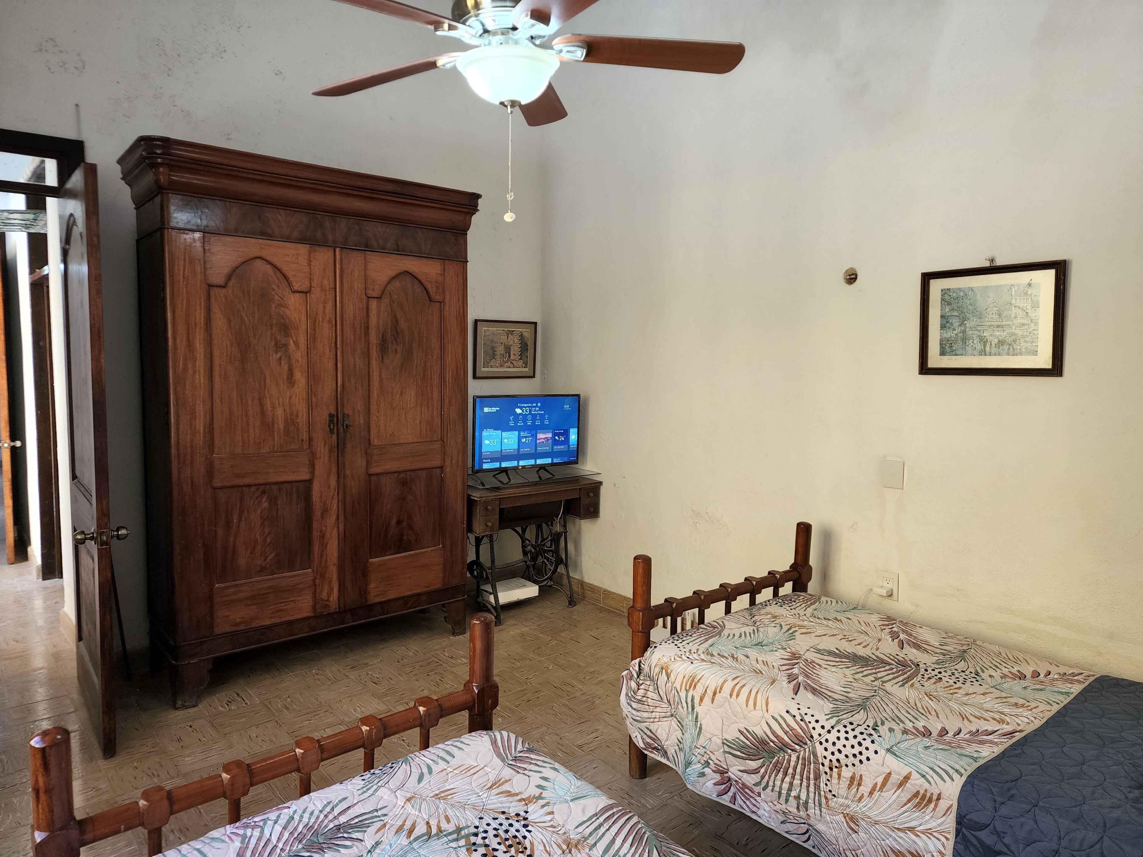 Un dormitorio con dos camas de madera, un armario de madera, un ventilador de techo, una máquina de coser y un televisor.