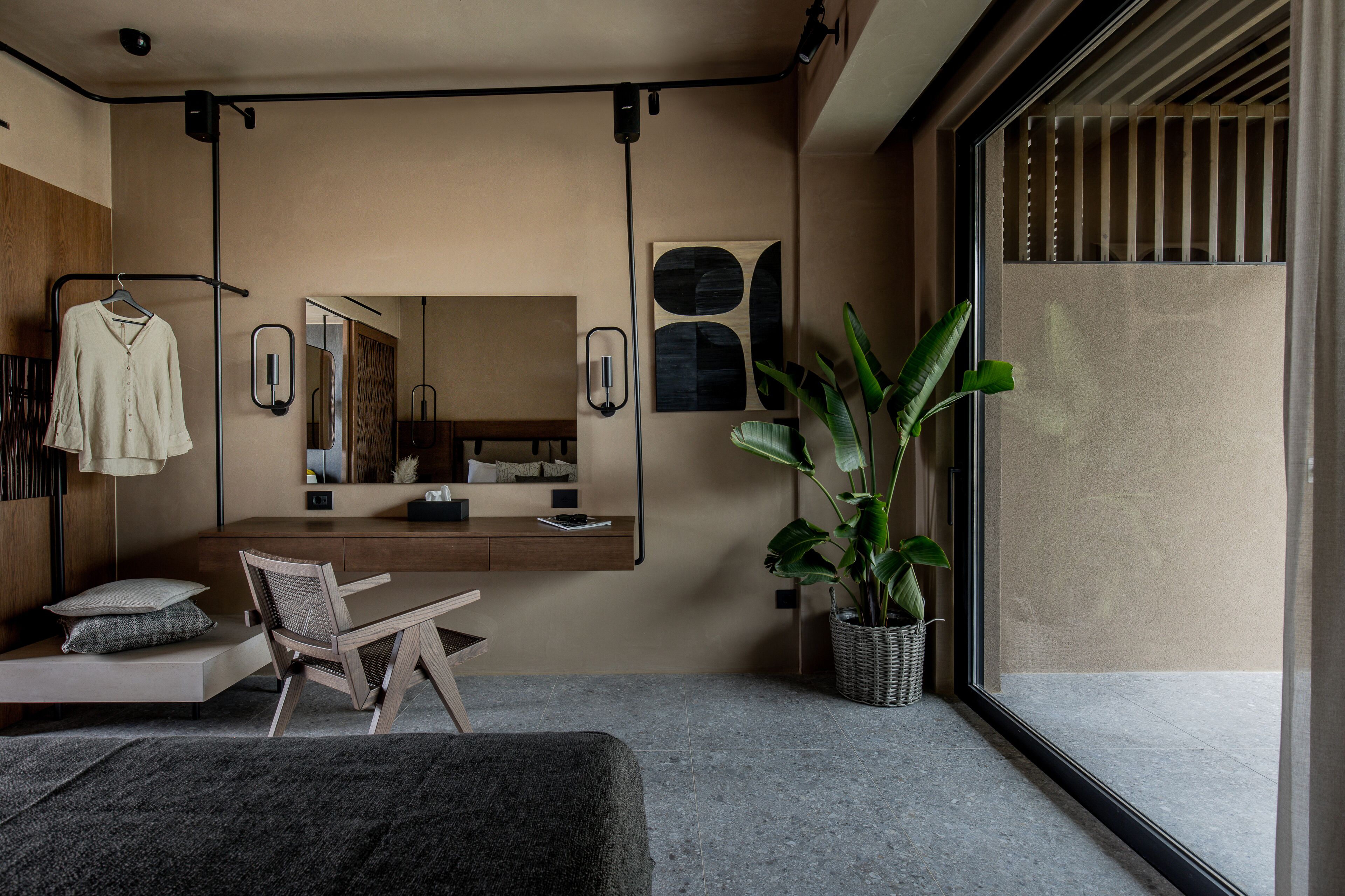 A modern bedroom with a large mirror, a bedside table, a chair, a hanging robe, and a potted plant.