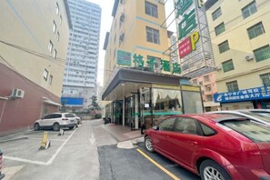 Entrada de un hotel con puerta de cristal, un coche rojo estacionado al frente y un edificio con varios pisos y ventanas.