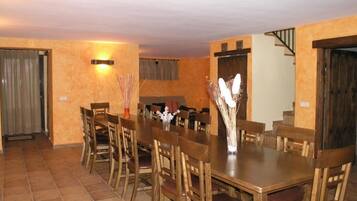 Un comedor con una larga mesa de madera y sillas, un florero con flores secas, y una escalera al fondo.