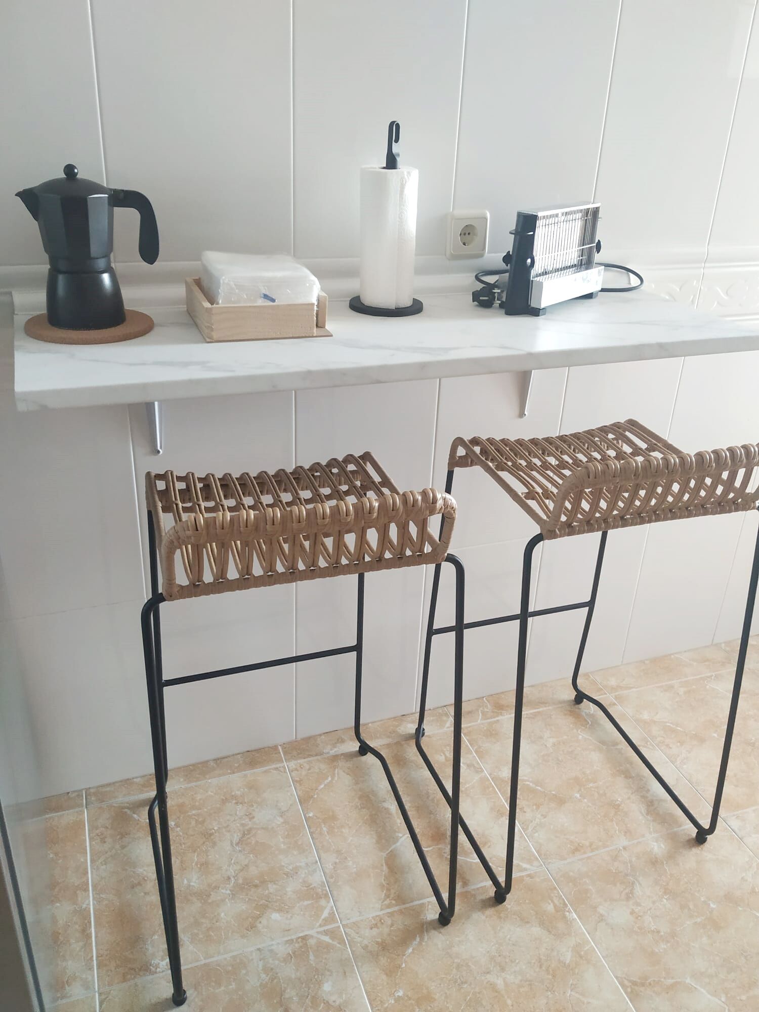Una cocina con dos taburetes de mimbre, una cafetera, una caja de pañuelos y una regleta en la encimera.