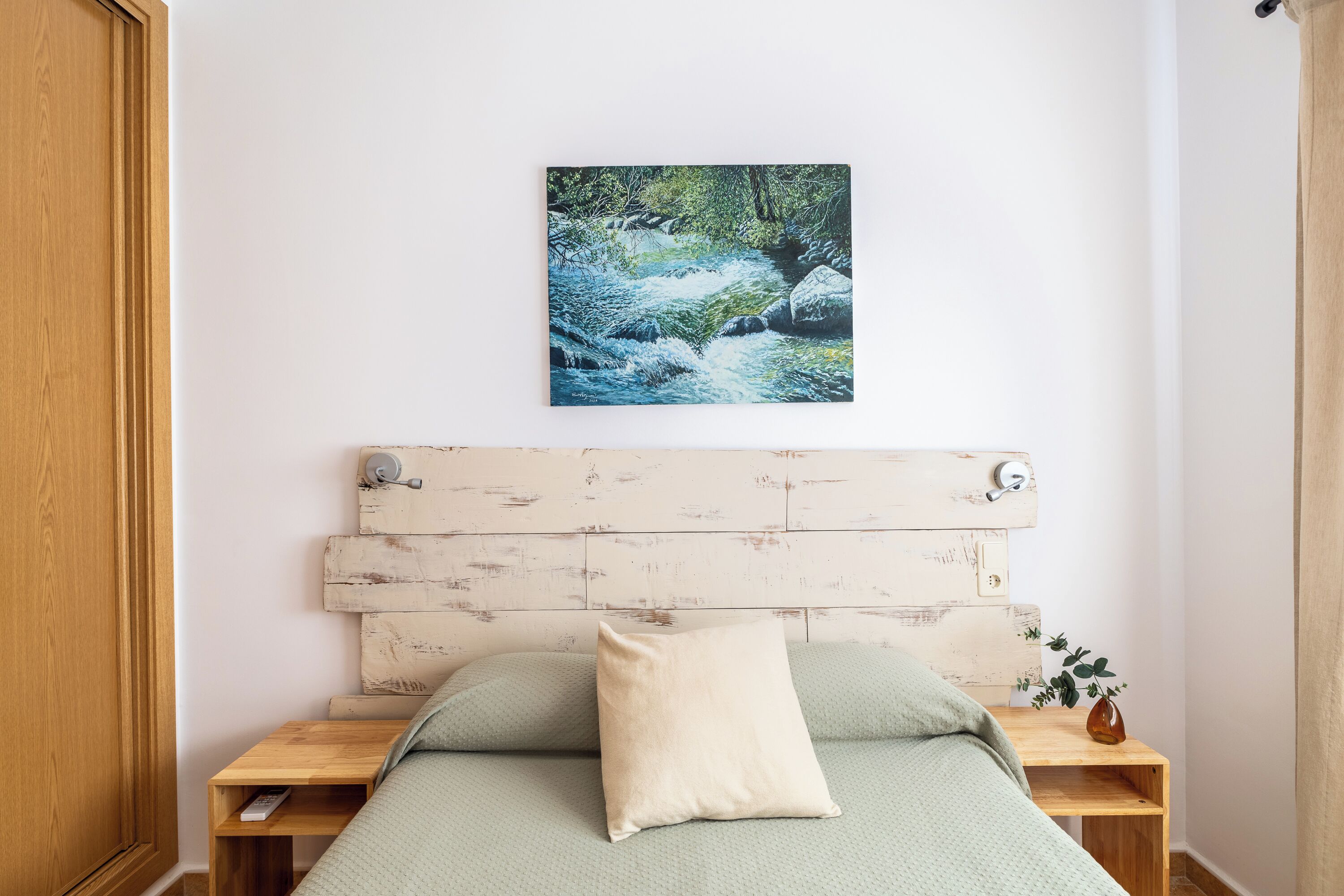 Un dormitorio con cabecera de madera, colcha verde, almohada color crema, una mesita de noche con un florero y un cuadro de un arroyo en la pared.