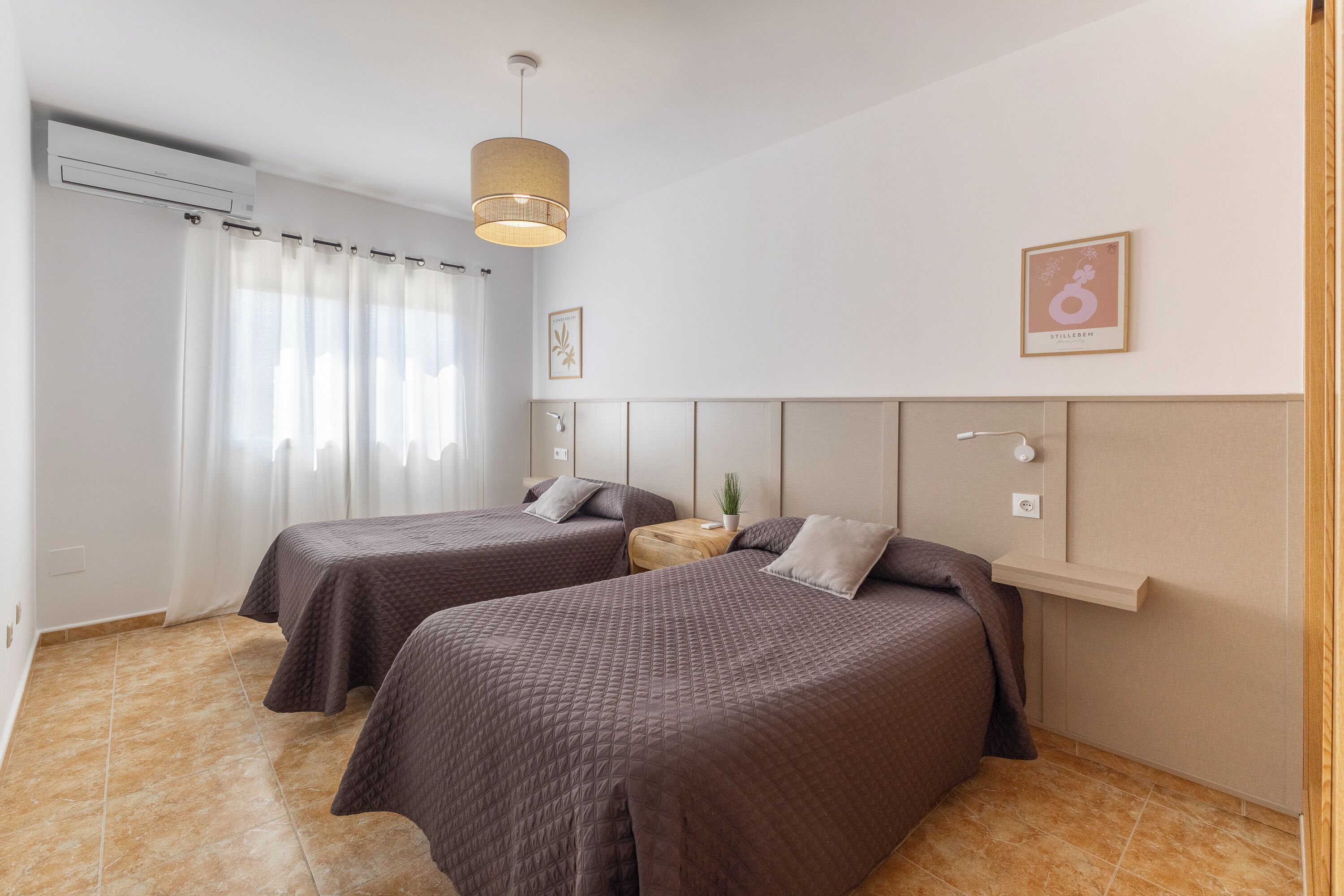 Ein Hotelzimmer mit zwei Betten, einem Holznachttisch, einer kleinen Topfpflanze und einem an der Wand befestigten Regal.