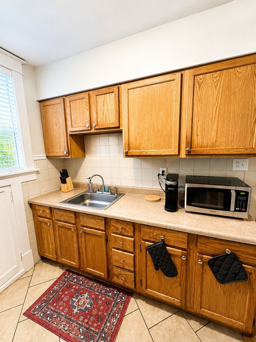 A kitchen with wooden cabinets, a sink, a kettle, a microwave, and a red patterned rug.
