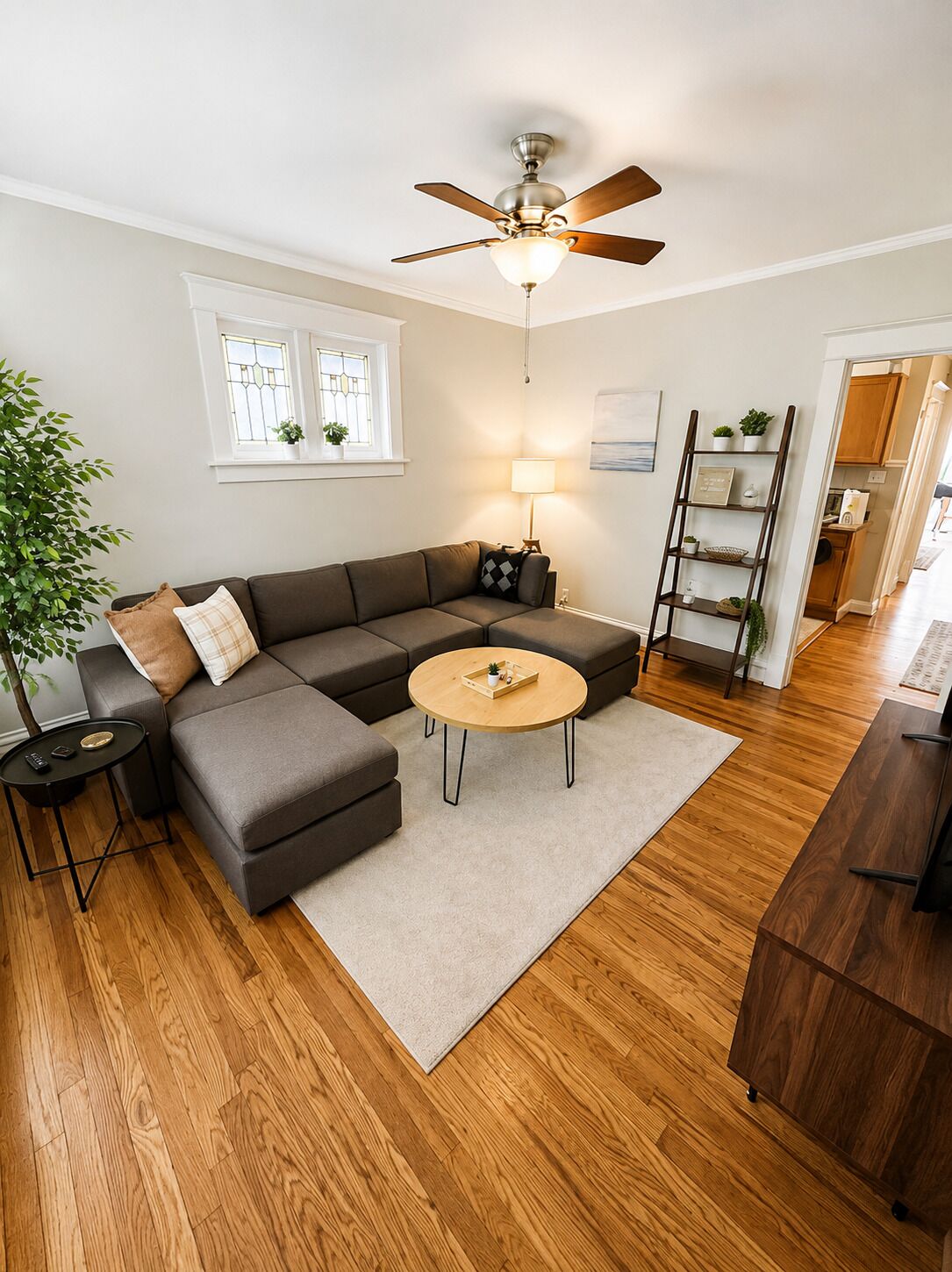 Un salon moderne avec un canapé d’angle, une table basse ronde, un ventilateur de plafond, une lampe, un plancher en bois et une fenêtre ornée de plantes.