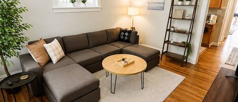 Un salon moderne avec un canapé d’angle, une table basse ronde, un ventilateur de plafond, une lampe, un plancher en bois et une fenêtre ornée de plantes.