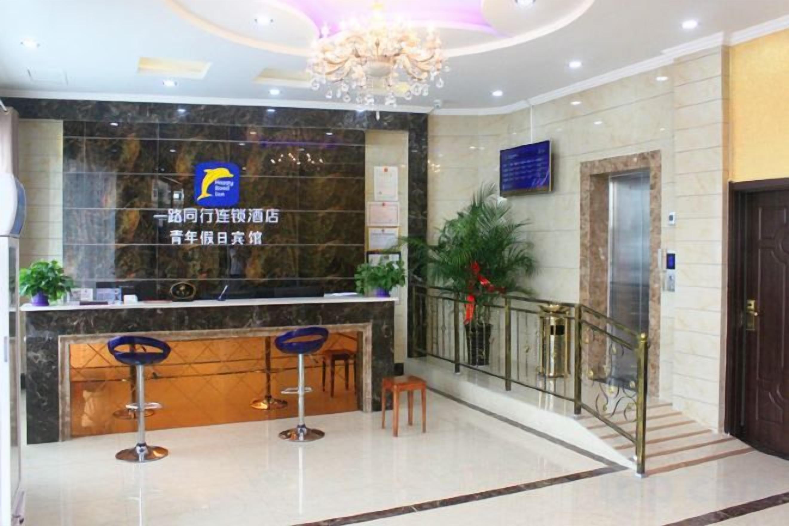 A hotel reception area with a marble counter, two bar stools, a chandelier, and a staircase with a metal railing.