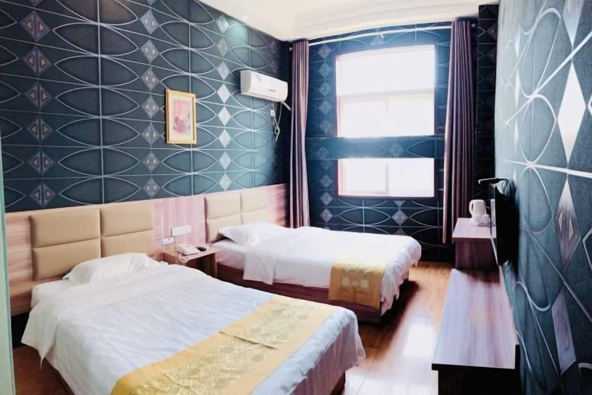 Una habitación de hotel con dos camas, una ventana con cortinas, una mesita de noche con lámpara y un aire acondicionado de pared.