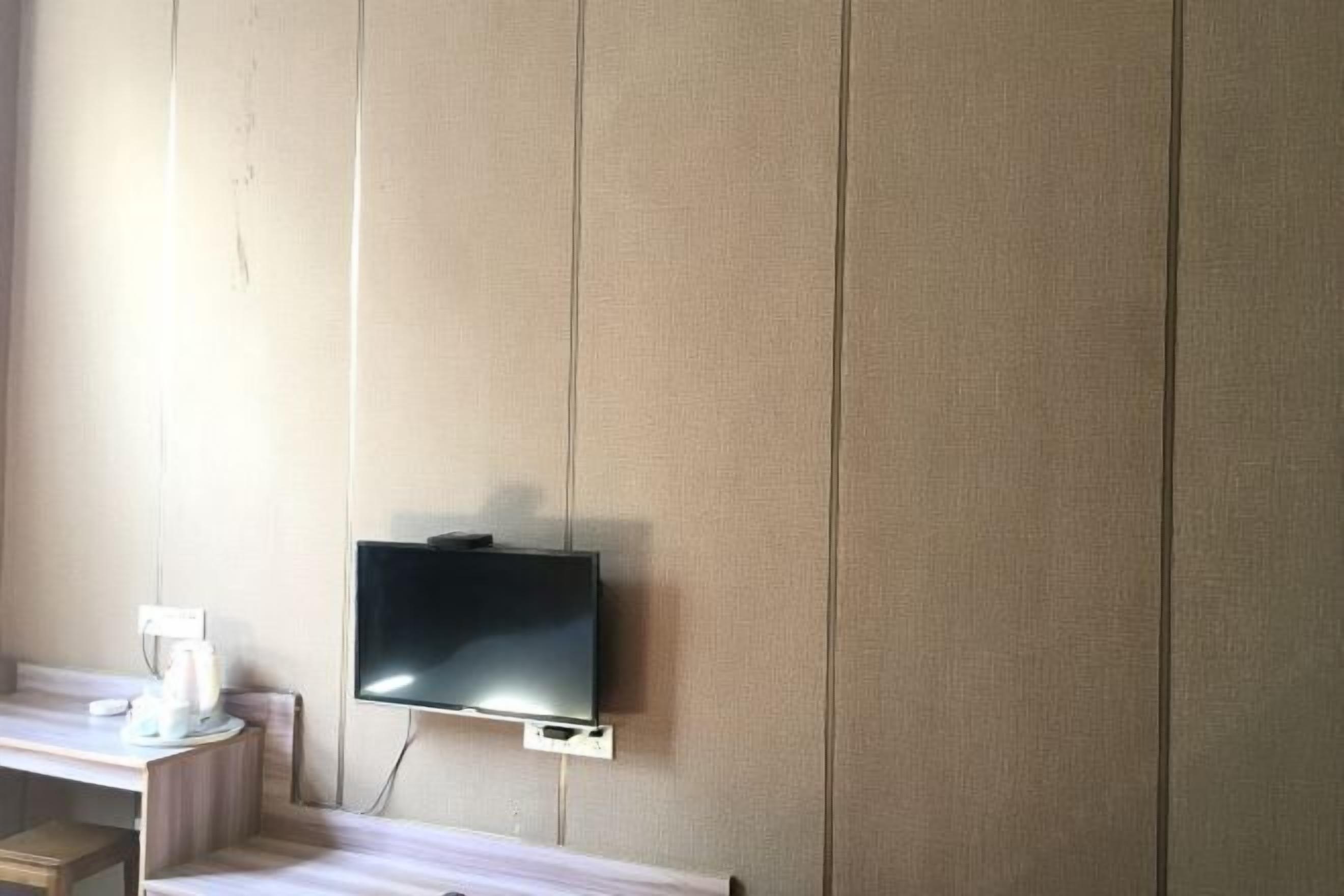 Una habitación con una televisión de pantalla plana montada en la pared, un escritorio de madera con una silla y un ventanal que deja entrar luz natural.
