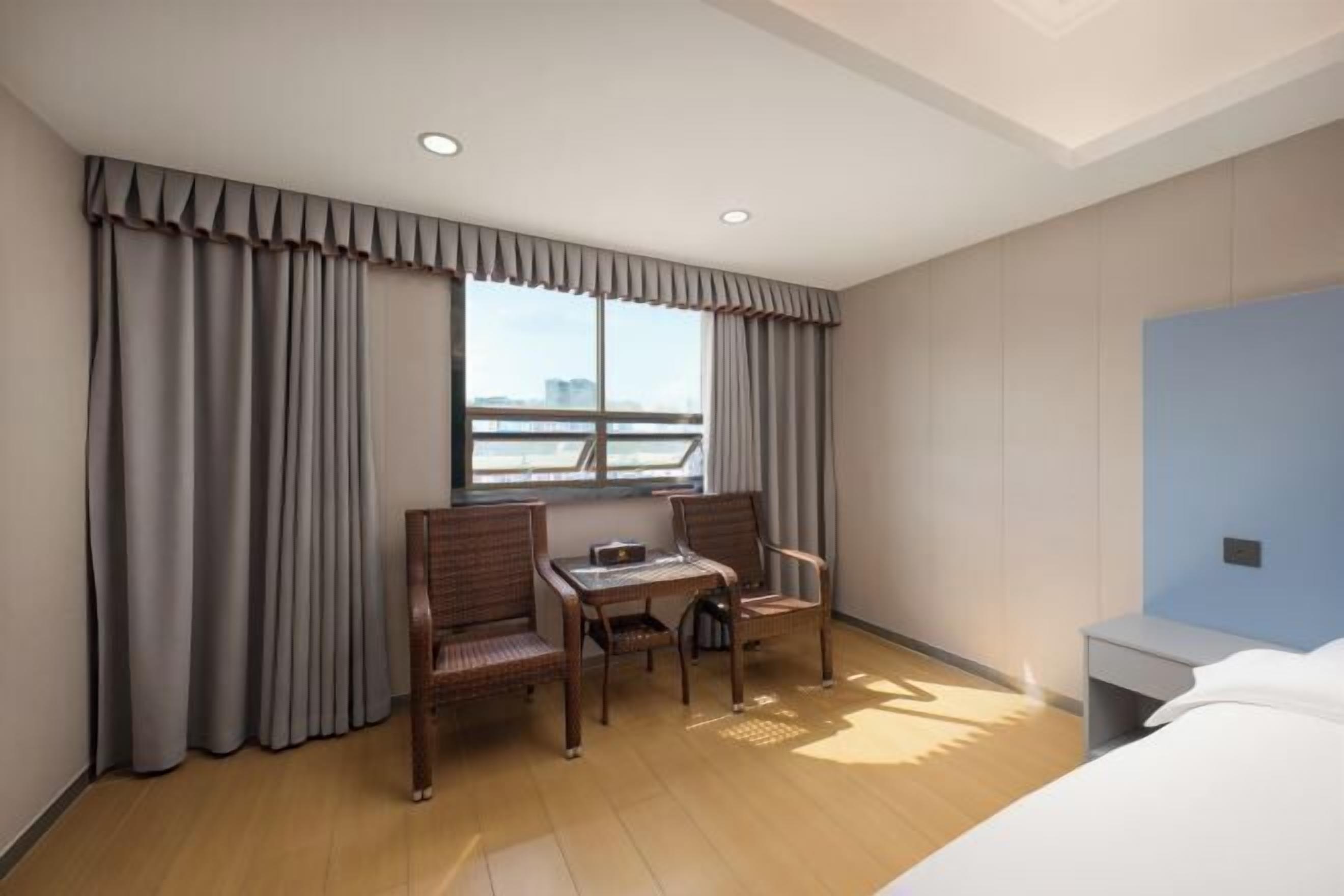 A hotel room with a bed, a desk, two chairs, a window with curtains, and a view of the city.