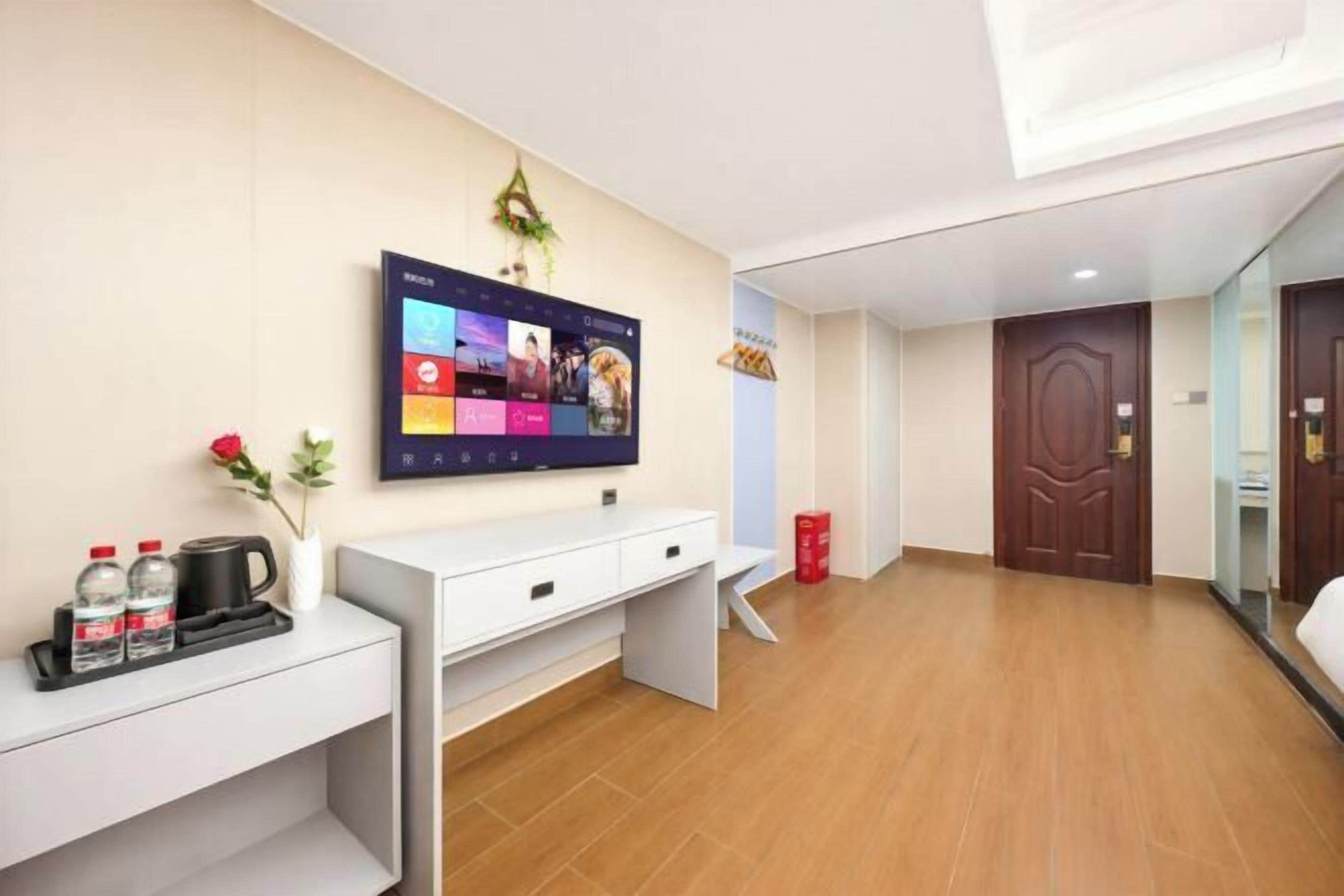 A hotel room with a flat-screen TV mounted on the wall, a white desk with a tray of bottled water and a coffee pot, a wooden floor, and a wooden door.