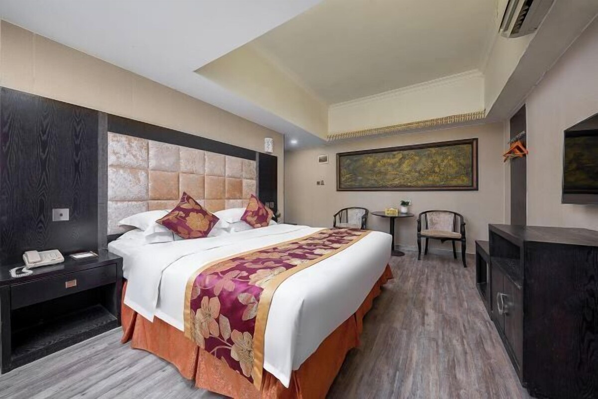 A hotel room with a large bed, a nightstand, a desk, a chair, and a painting on the wall.