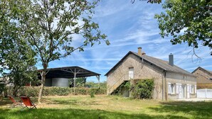 Ein rustikales Bauernhaus mit überdachtem Außenbereich, umgeben von üppiger Vegetation und blauem Himmel.