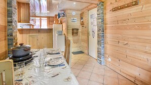 A wooden interior with a dining table set for four, a refrigerator, and a kitchen area with wooden cabinets and a stone wall. - Newly Renovated 2BR Apartment Near Mont Chéry Cable Car - Les Gets, Sleeps 5 (Les Gets)