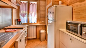 A compact kitchen with wooden cabinets, a refrigerator, a microwave, and a window with curtains. - Newly Renovated 2BR Apartment Near Mont Chéry Cable Car - Les Gets, Sleeps 5 (Les Gets)