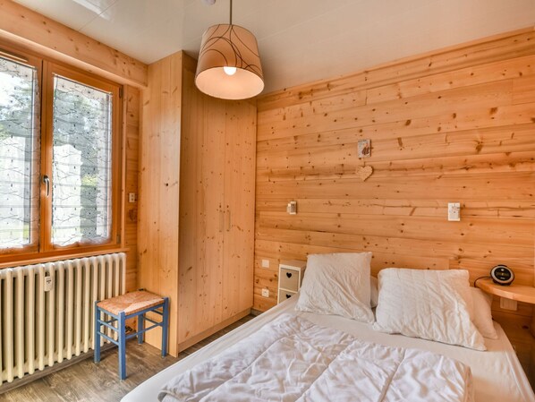 A wooden cabin bedroom with a bed, a small chair, a window, and a wall-mounted heater. - Newly Renovated 2BR Apartment Near Mont Chéry Cable Car - Les Gets, Sleeps 5 (Les Gets)