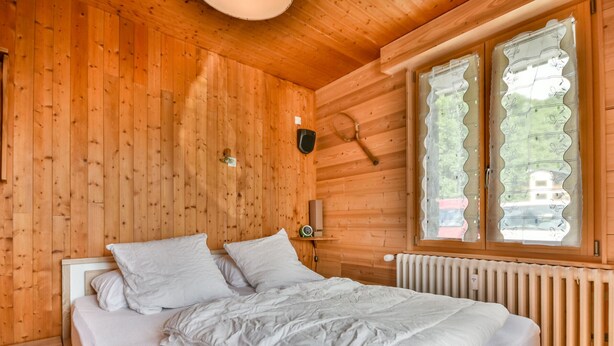 A wooden cabin bedroom with a bed, a nightstand, a wall clock, and a window with decorative curtains. - Newly Renovated 2BR Apartment Near Mont Chéry Cable Car - Les Gets, Sleeps 5 (Les Gets)