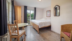 Una habitación con una cama, una mesa con sillas, un sillón de mimbre, una ventana con cortinas y un espejo en la pared.