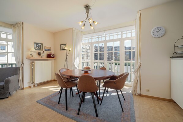 Un coin repas avec une table ronde, six chaises, un lustre, un radiateur, un canapé et une vue sur les bâtiments à travers de grandes fenêtres.