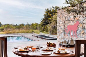 A dining table set with plates, glasses, and a fruit basket, overlooking a swimming pool and a stone wall with a horse silhouette. - Bijapur Lodge (Bali)