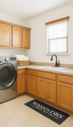 A compact kitchen with wooden cabinets, a washing machine, a sink, and a window with blinds. - Kimberly's Private Palm Château Salt Pool & Hot Tub (Lake Elsinore)